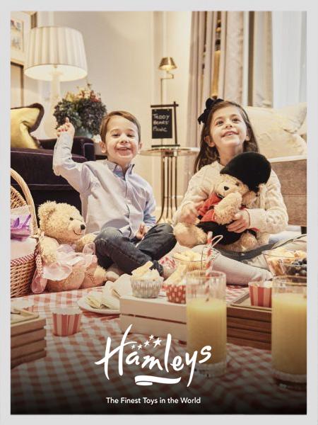 Two happy children enjoying an indoor teddy bear picnic with Hamleys toys and stuffed animals.